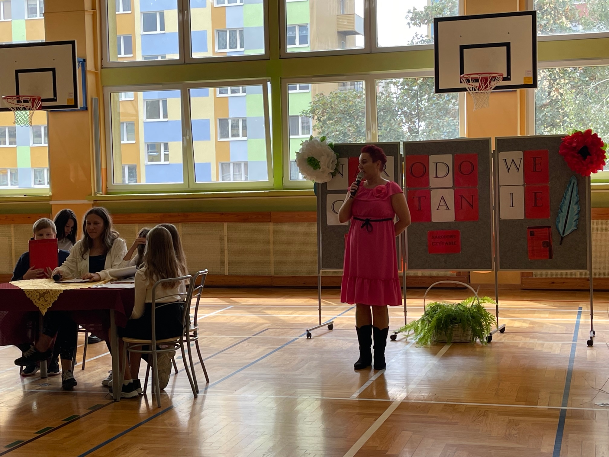 Bibliotekarka szkoły na dużej sali gimnastycznej wygłasza przemówienie dotyczące narodowego czytania.